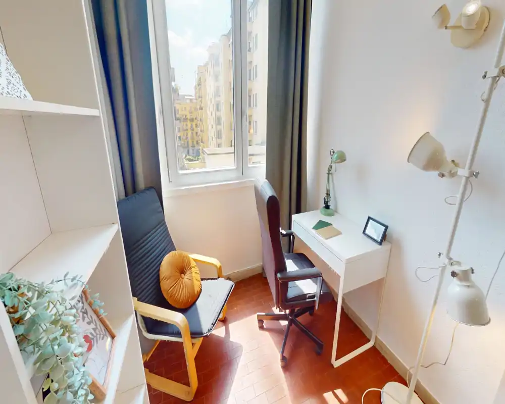 Angolo giorno/studio luminoso e accogliente con finestra che illumina la stanza, una poltrona con cuscino decorativo, una scrivania con lampada e una sedia da ufficio — ideale per leggere o lavorare da casa.