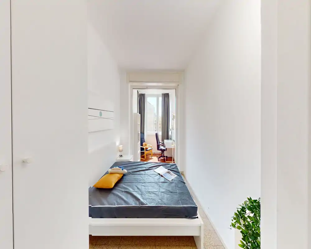 Camera da letto luminosa e stretta con letto matrimoniale in primo piano e una postazione vicino alla finestra visibile attraverso la porta; arredo moderno e pochi dettagli caldi.