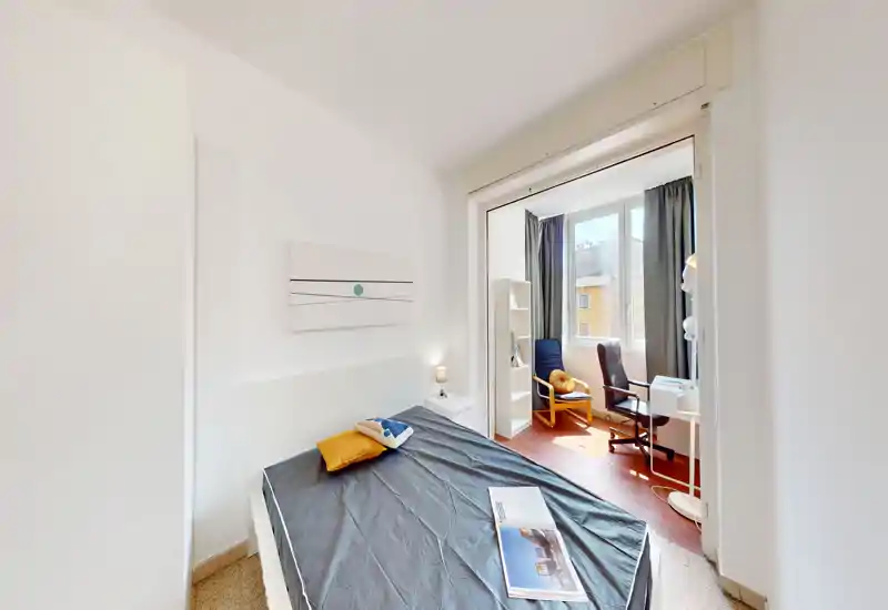 Camera da letto moderna e luminosa con letto matrimoniale, comodino con lampada e un piccolo angolo studio vicino alla finestra soleggiata. Pareti bianche e arredo essenziale creano un'atmosfera accogliente e pulita.