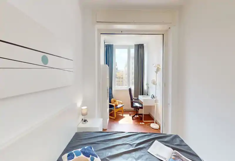 Camera da letto luminosa e minimal con letto ben visibile in primo piano e una postazione di lavoro soleggiata vicino alla finestra. Pareti bianche, una lampada da comodino e un cuscino decorativo creano un’atmosfera moderna e rilassante.