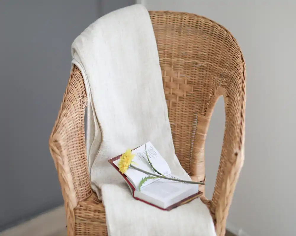 Rincón acogedor con un sillón de mimbre cubierto por una manta clara y un libro abierto con una flor amarilla: un tranquilo espacio de lectura con texturas naturales y luz suave.
