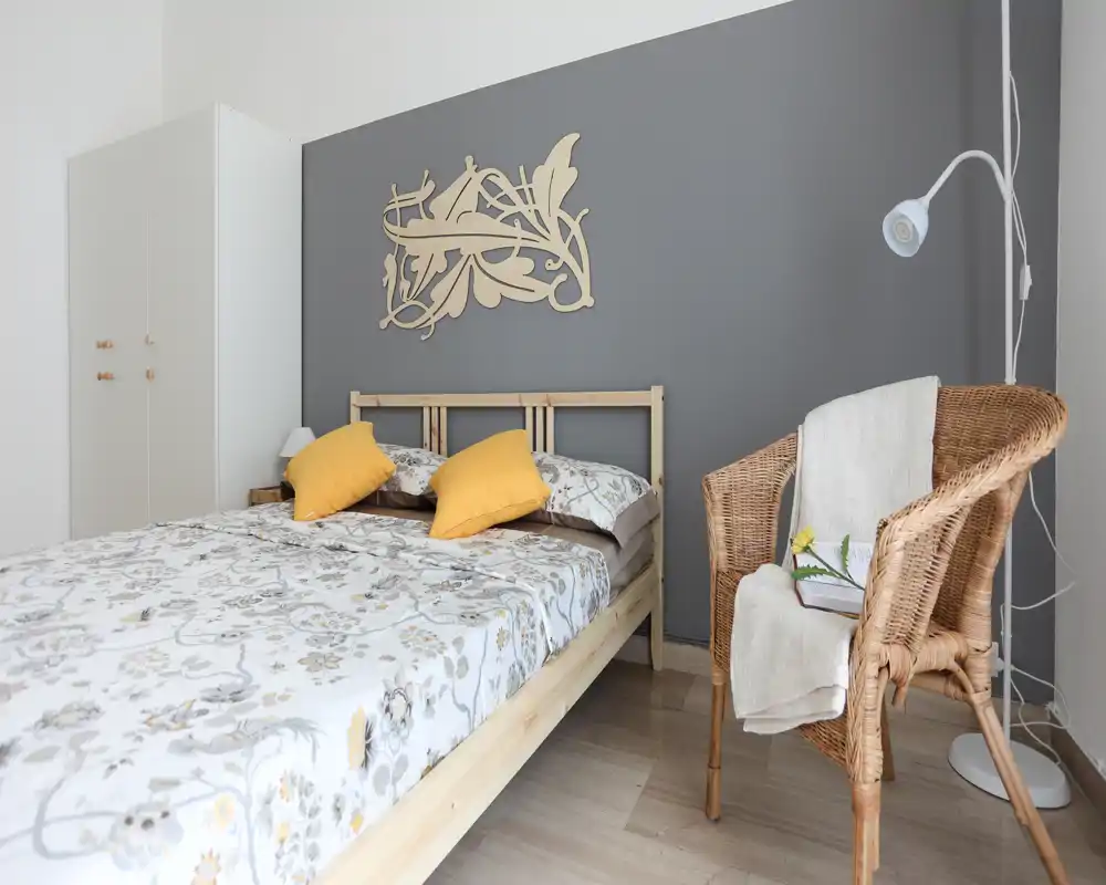 Dormitorio luminoso y bien decorado con cama doble de madera, ropa de cama estampada y cojines amarillos. Una silla de mimbre y un armario ordenado completan el espacio acogedor y moderno.