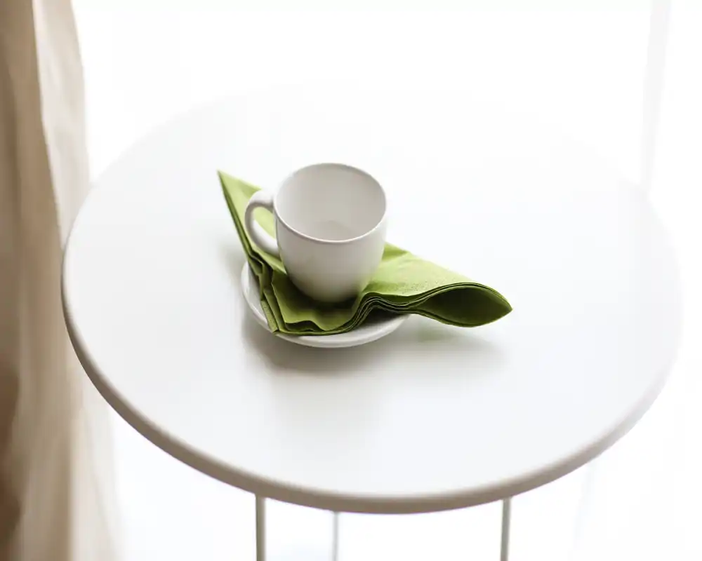 Mesa redonda minimalista con una taza y servilleta verde junto a una ventana luminosa: un rincón de café sencillo, limpio y luminoso.