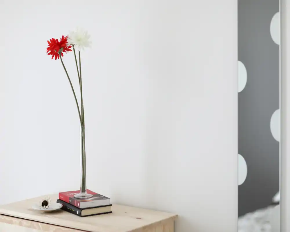Rincón decorativo minimalista con un jarrón de vidrio alto con dos flores de tallo largo sobre una superficie de madera; en el borde derecho se ve parcialmente un espejo o panel con diseño.