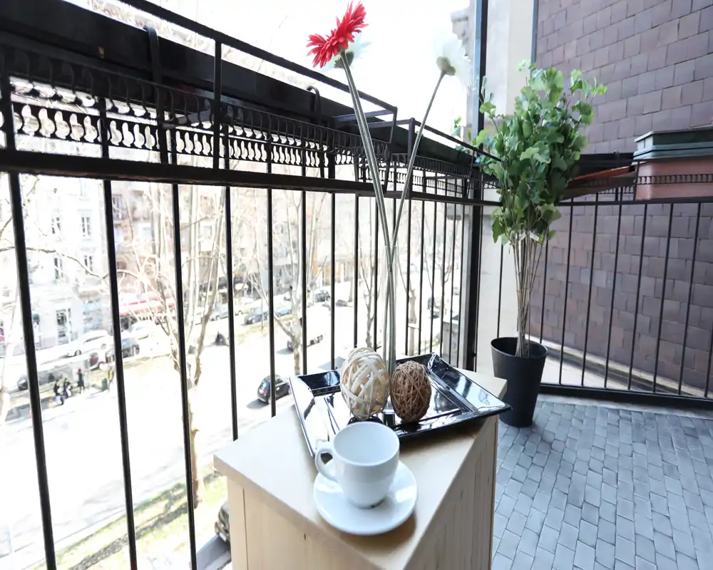 Balcón soleado con una pequeña mesa, bandeja decorativa y una taza, con vistas a una calle arbolada — ideal para el café de la mañana.