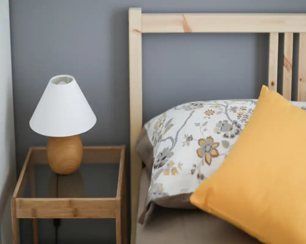 Primer plano de una esquina de dormitorio con cabecero de madera, almohada con estampado y cojín amarillo junto a una mesita con lámpara — luminoso, acogedor y bien decorado.