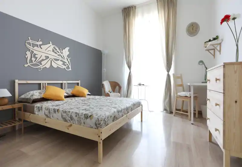 Dormitorio luminoso y moderno con cama doble de madera, ropa de cama con estampado y cojines amarillos; gran ventana con cortinas que ofrece abundante luz natural y un acogedor rincón de lectura.