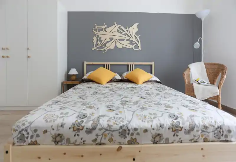 Dormitorio luminoso y bien decorado con una gran cama de madera, ropa de cama floral y cojines amarillos; pared gris y sillón de mimbre crean un ambiente acogedor.