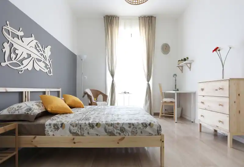 Dormitorio luminoso y aireado con cama doble de madera con ropa de cama floral y cojines mostaza, cómoda y un pequeño escritorio junto a la ventana.