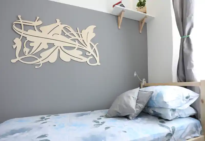 Dormitorio individual luminoso en esquina con cama arreglada, ropa de cama azul floral, cojines decorativos y adorno en la pared: espacio compacto y acogedor con luz natural.