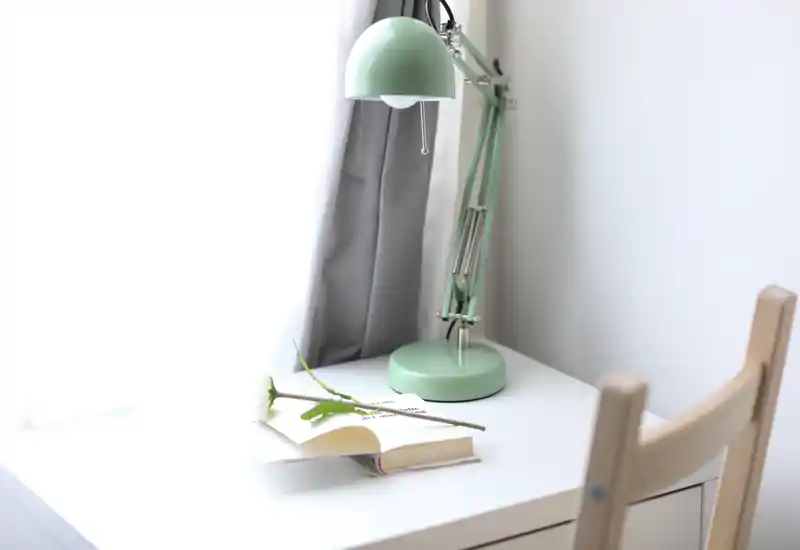 Esquina luminosa de mesita/escritorio con lámpara ajustable verde menta, libro abierto y una flor sobre una superficie blanca — ideal como rincón de lectura o estudio en una habitación.