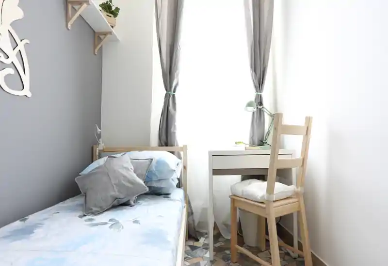Dormitorio pequeño y luminoso con cama individual de madera, ropa de cama azul claro con motivos florales, cojines decorativos y un escritorio junto a la ventana — acogedor y bien decorado.