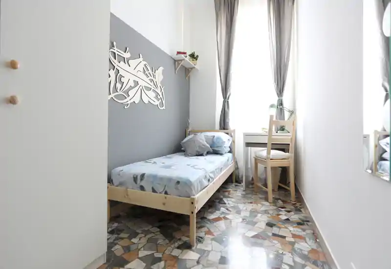 Habitación individual luminosa y compacta con cama de madera, panel decorativo en la pared y un pequeño escritorio junto a la ventana: ideal para estudiante o viajero solo.