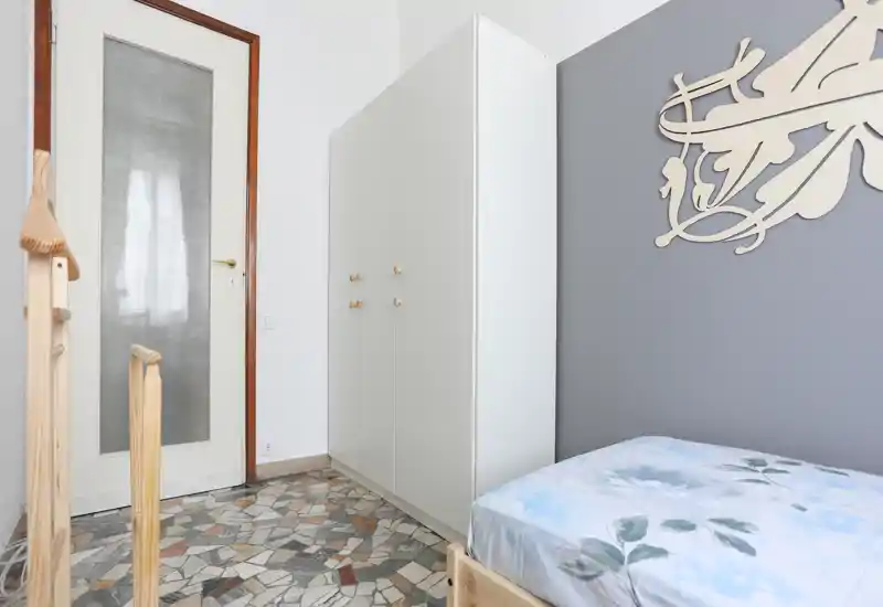 Rincón de dormitorio luminoso con cama individual, armario blanco y decoración en la pared; suelo de mosaico y luz natural ofrecen una sensación fresca.