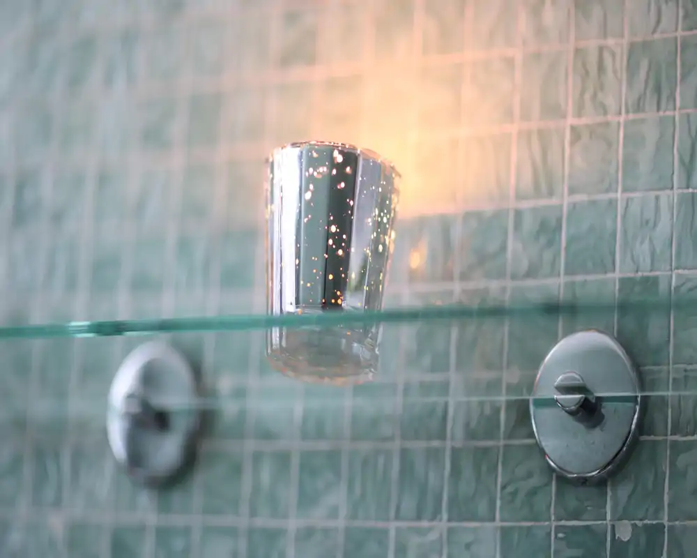 Plano detalle de pared de baño con mosaico, una repisa de vidrio y un pequeño vaso metálico decorativo; buena nitidez para destacar acabados y accesorios.