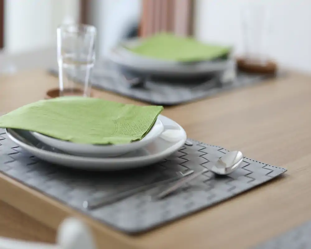 Primer plano de una mesa puesta con platos, servilletas verdes y cubiertos sobre individual — una zona de comedor acogedora con luz natural suave.