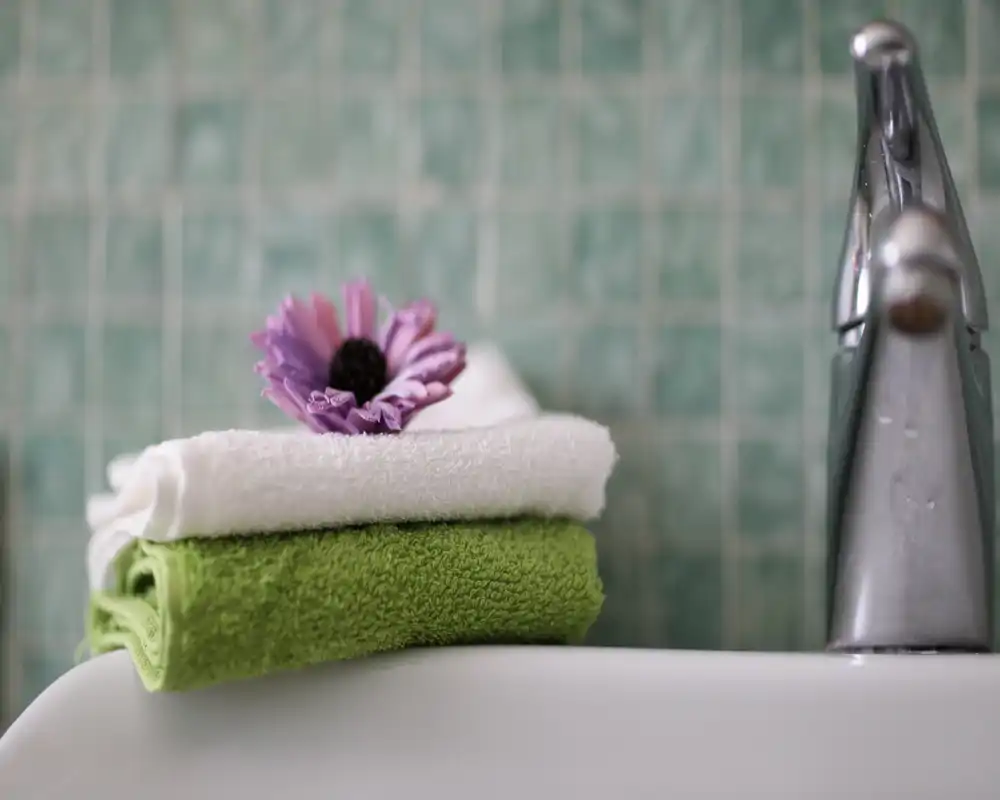 Primer plano del lavabo del baño con toallas blancas y verdes dobladas, una flor morada decorativa y un grifo cromado frente a un revestimiento de mosaico verde.
