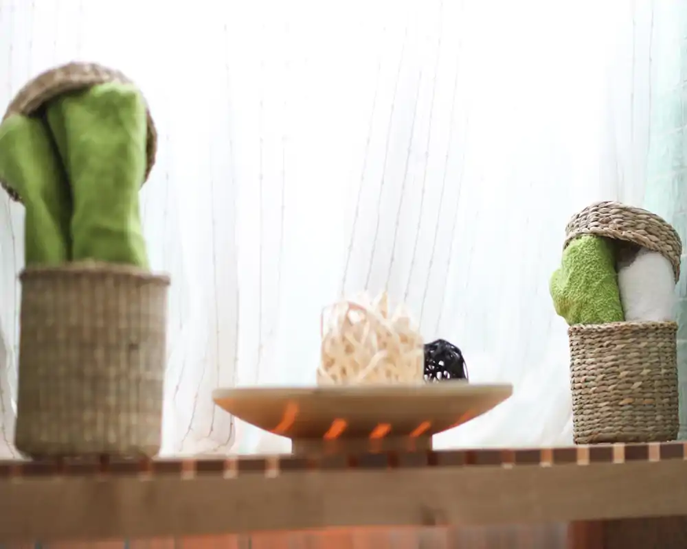 Vigneta decorativa de estilo exterior con cestas trenzadas y toallas enrolladas sobre una superficie de madera frente a cortinas translúcidas; luz natural suave que crea un rincón relajante tipo spa.