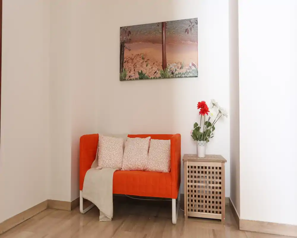 Rincón de estar luminoso y bien decorado con un sofá pequeño naranja con cojines, mesita y flores decorativas — ideal para leer o relajarse.