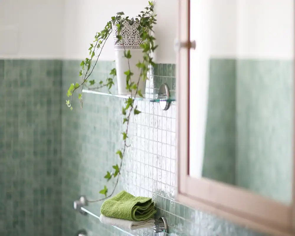 Rincón de baño luminoso con azulejos mosaico verdes, estantes de vidrio con toallas dobladas y una planta colgante junto a un armario con espejo.