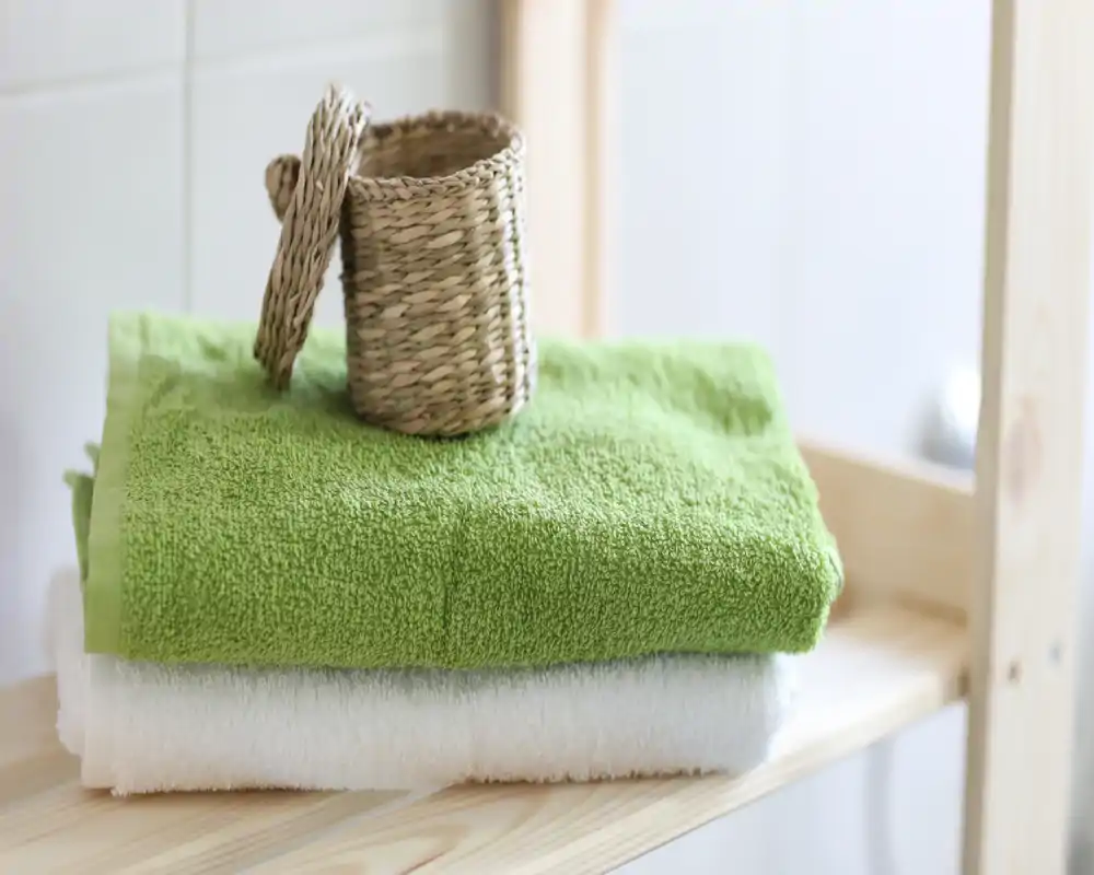 Primer plano de toallas verdes y blancas dobladas en una estantería de madera con una pequeña cesta trenzada — imagen luminosa, limpia y de estilo minimalista, ideal para anunciar un baño.