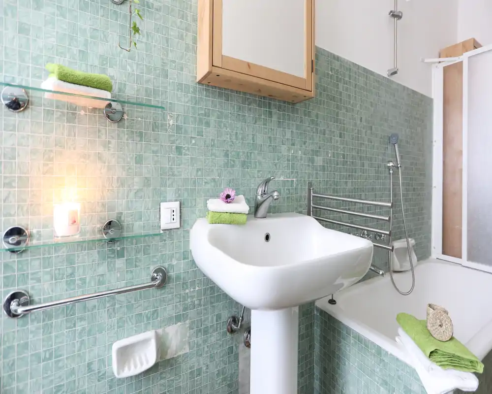 Baño luminoso con lavabo de pedestal blanco, bañera con ducha de mano, azulejos de mosaico verde, toallas a juego y un pequeño armario con espejo.