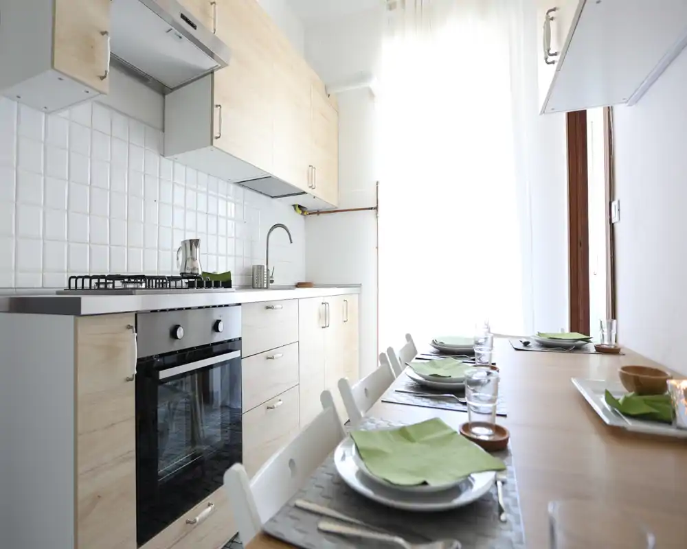 Cocina moderna y luminosa en línea con muebles de madera clara, horno empotrado y mesa puesta para cuatro — espacio compacto y con estilo, muy luminoso.