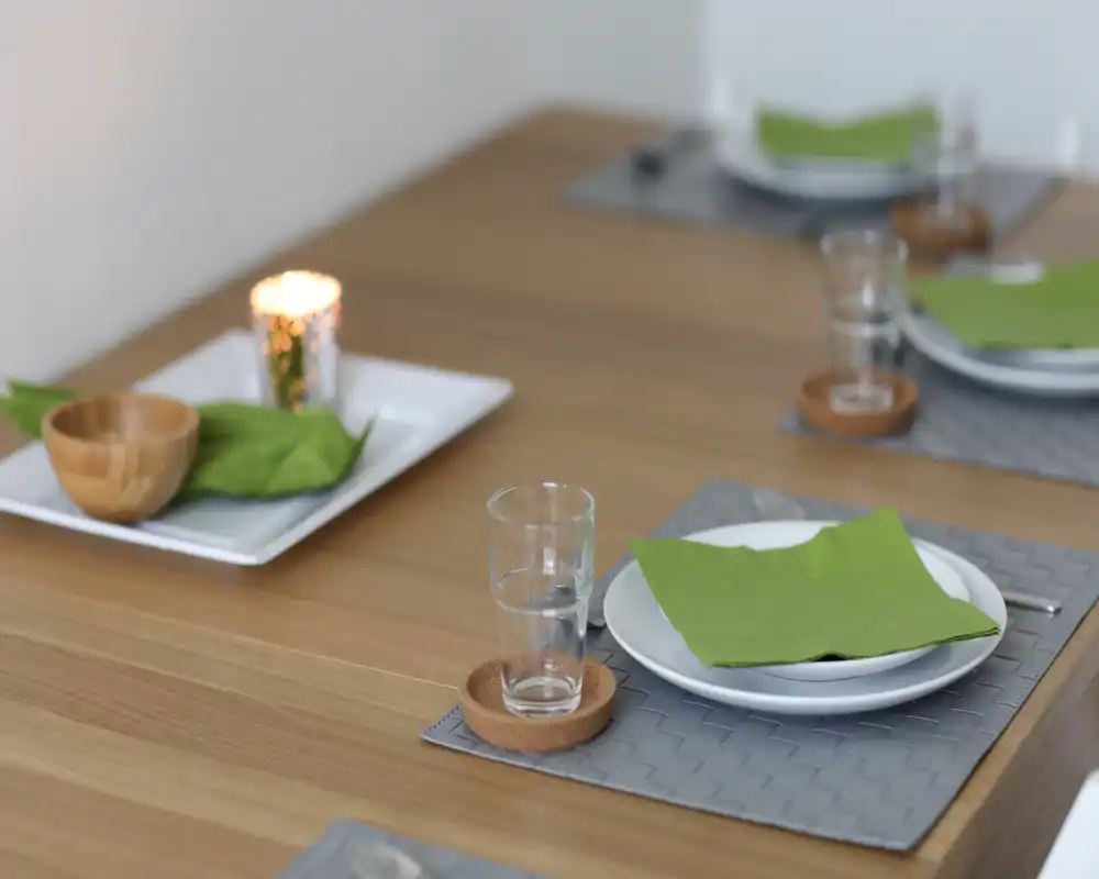 Primer plano de una mesa puesta con platos, vasos, servilletas verdes y una vela sobre madera: un comedor estilizado y acogedor listo para comer.