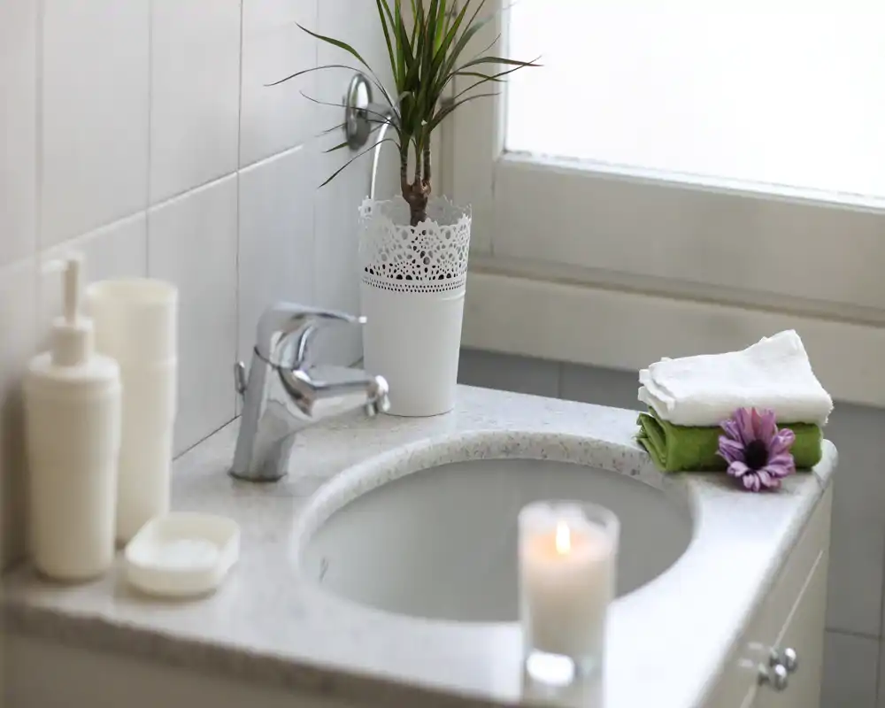 Zona del lavabo del baño luminosa y limpia con grifo cromado moderno, dispensadores de jabón, toallas dobladas y una pequeña planta decorativa y vela para un toque acogedor.