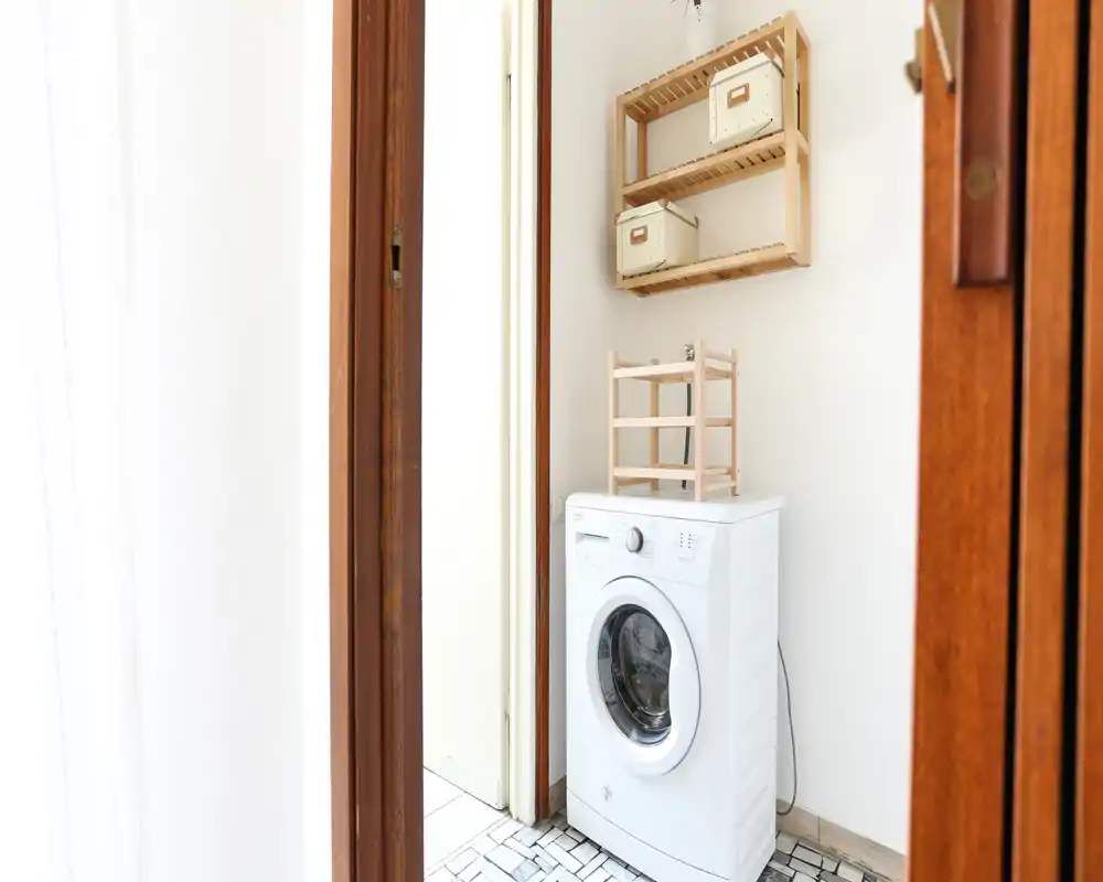 Pequeño rincón de lavandería luminoso con lavadora de carga frontal, estantes en la pared y suelo de baldosas ordenado — espacio de servicio funcional.