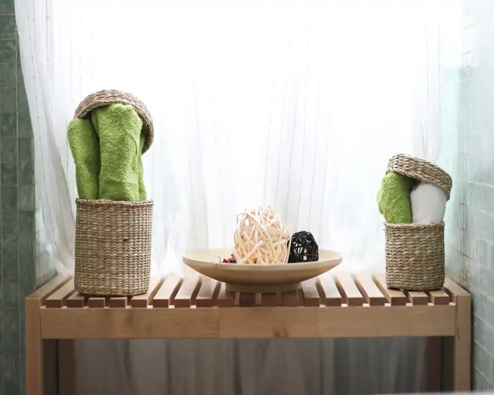 Exhibición de baño ordenada con cestas tejidas que contienen toallas enrolladas y un cuenco decorativo sobre un banco de lamas de madera; suave luz natural a través de cortinas translúcidas.