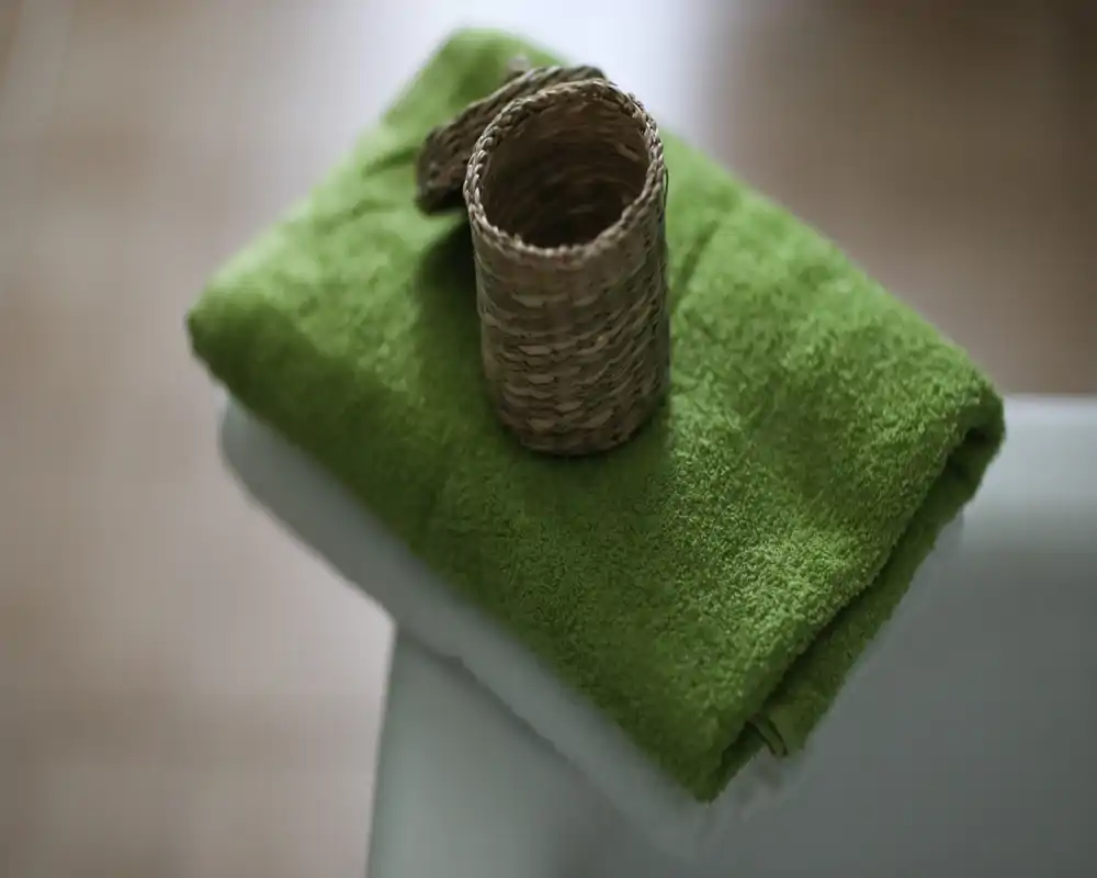Primer plano de toallas dobladas verdes y blancas con una pequeña cesta tejida sobre una superficie neutra. La imagen resalta la textura y el estilo, adecuada para mostrar ropa de cama o amenities.