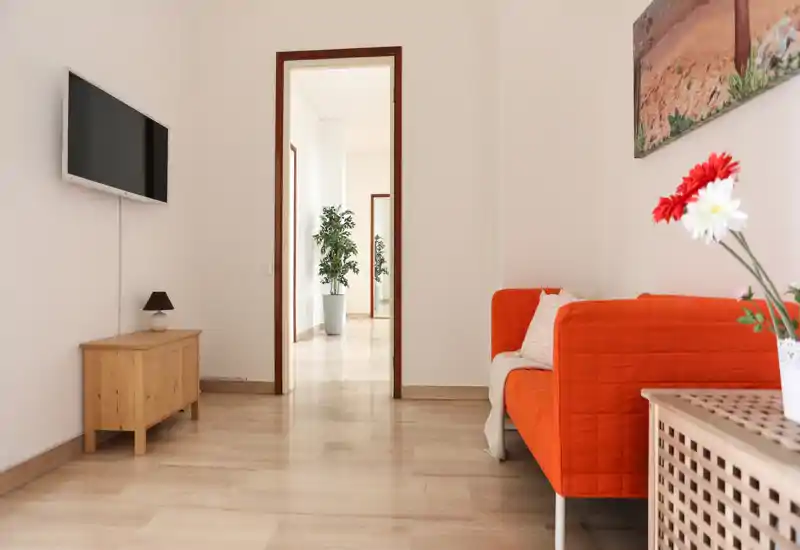 Salon lumineux et minimaliste avec un canapé orange, une TV murale et une lumière naturelle créant une ambiance accueillante.