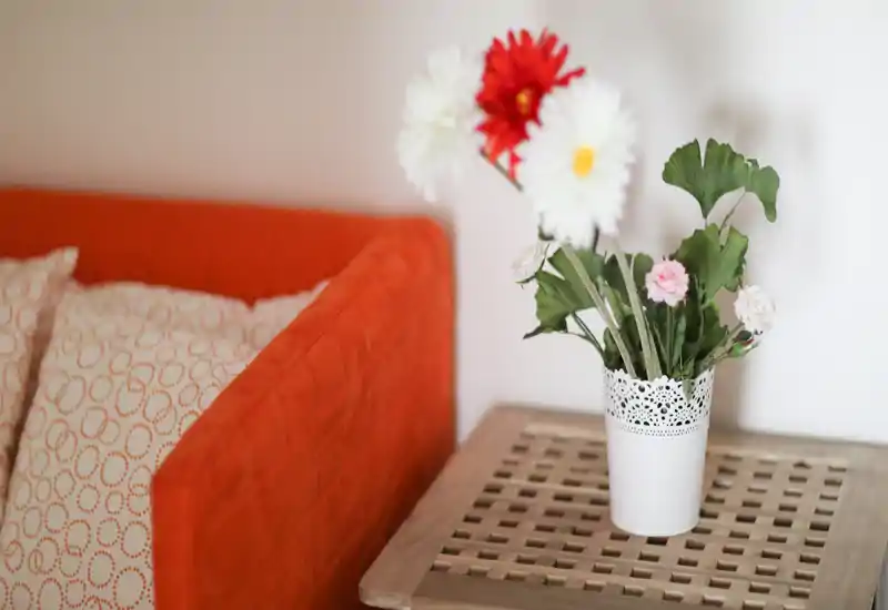 Détail d'un salon chaleureux avec un canapé orange, des coussins à motifs et un vase de fleurs décoratif sur une table d'appoint en bois — lumière douce et ambiance accueillante.