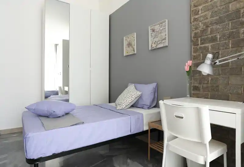 Neat single bedroom with a comfortable bed dressed in light lilac linens, a white desk and chair beside a textured brick accent wall — bright, modern and well styled.