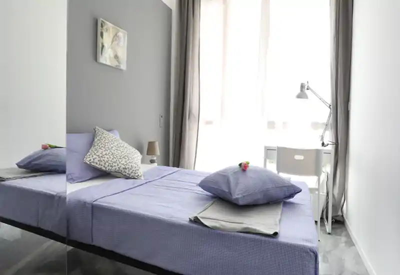 Bright, modern bedroom with a neatly made single/double bed dressed in soft lilac linens, decorative cushions, and a bedside lamp; a desk by the window creates a practical work corner.