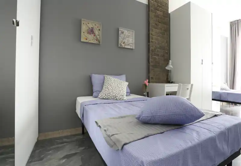 Bright, modern bedroom with a single bed dressed in soft lilac linens, decorative pillows, a small desk and wardrobe; neutral gray accent wall and exposed brick detail create a stylish, cozy feel.