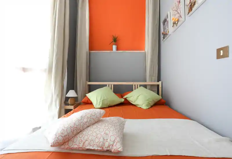 Bright, cozy bedroom with an orange bedspread, decorative pillows and soft natural light from a large window — ideal for rental photos showcasing comfort and style.