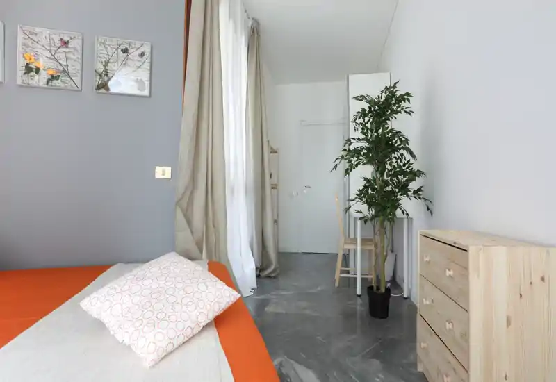 Bright, modern bedroom with an orange-accent bed, soft textiles and natural light from a tall window; simple wooden storage and a potted plant add a fresh, minimal touch.