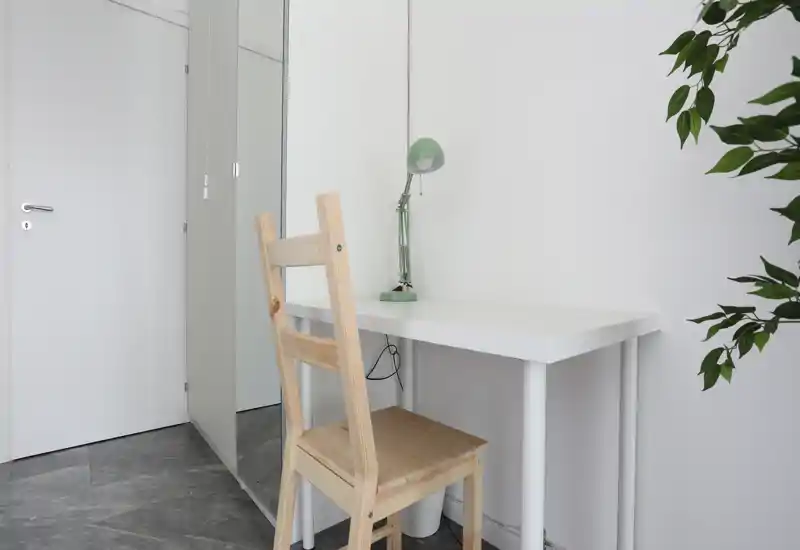 Minimal living area with a small white desk, wooden chair and a green desk lamp beside a potted plant — a compact home office corner in a neutral, bright space.