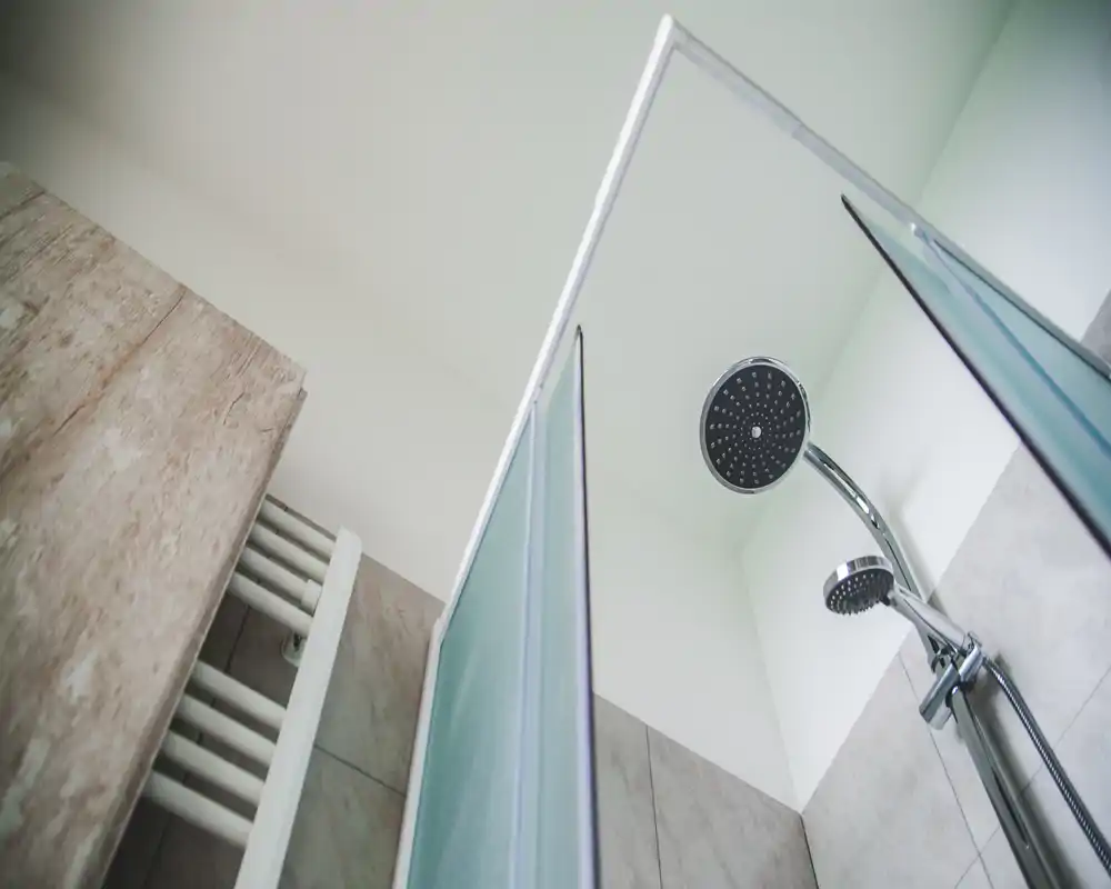 Modern shower corner with a chrome rain showerhead and handheld spray, frosted glass door and tiled walls — bright, minimal bathroom detail.