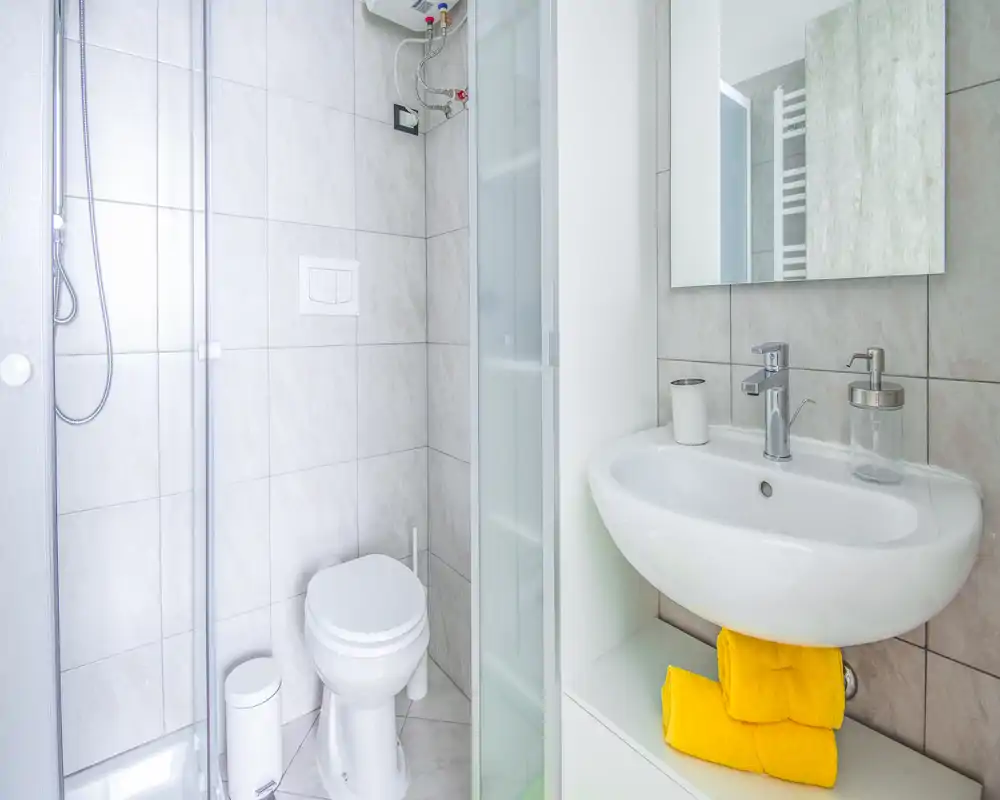 Compact, modern bathroom with a corner shower, wall-mounted toilet and a white sink. Bright, clean tiles and cheerful yellow towels create a fresh, inviting feel.