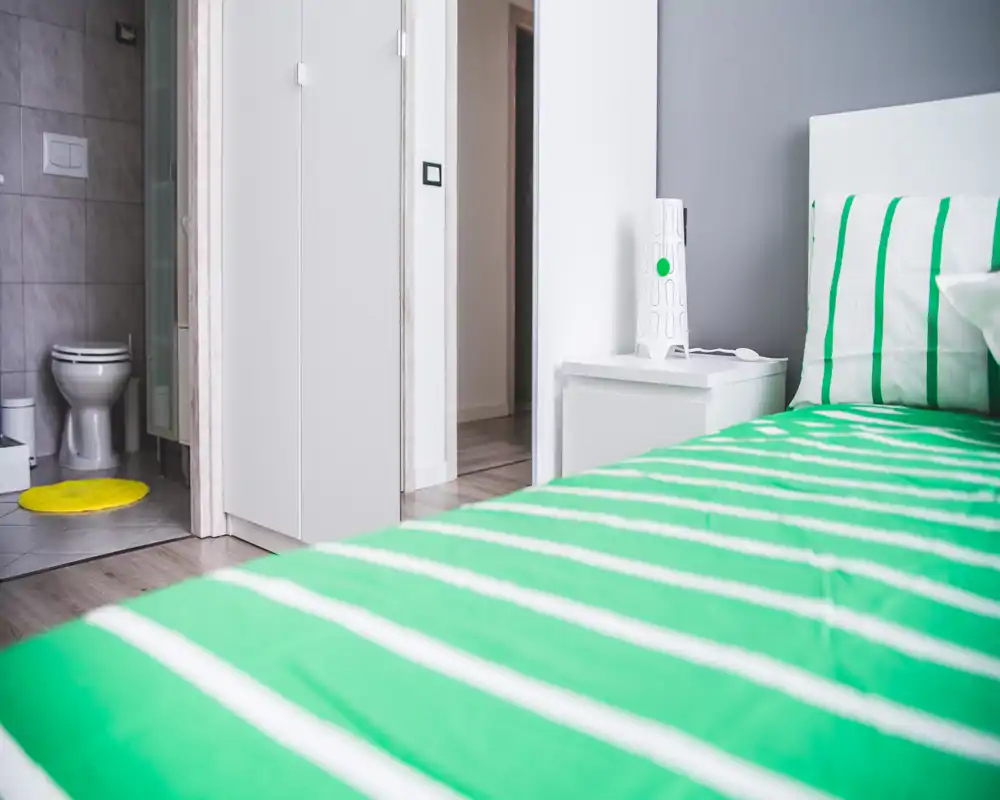 Bright modern bedroom corner with a green-striped bed in the foreground, a white bedside table and lamp, and an en-suite bathroom visible through an open door.