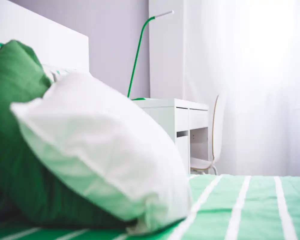 Bright, modern bedroom corner with a green-striped bed, soft pillows and a small white desk by a sunlit window — a cozy, well-lit space ideal for rest or light work.