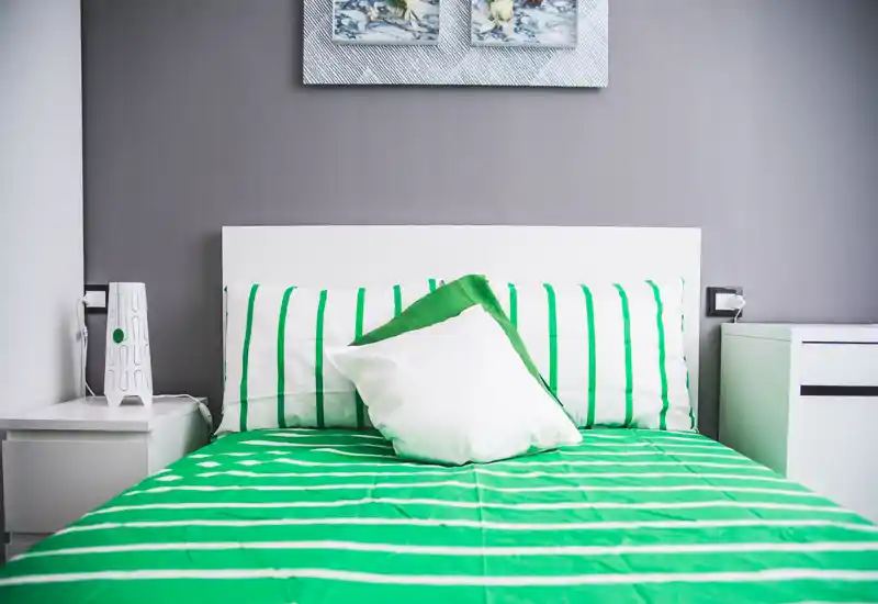 Bright, modern bedroom with a neatly made double bed in green-and-white striped linens, matching pillows and minimalist bedside tables — clean, fresh and inviting for guests.