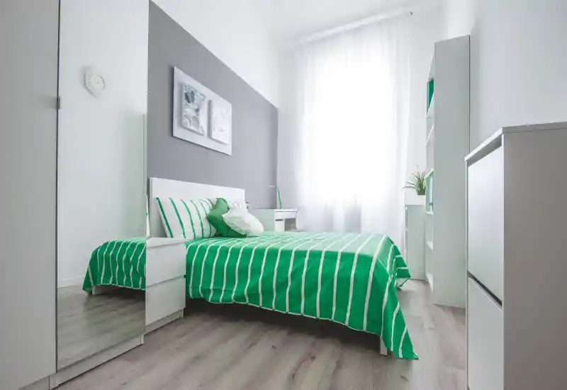 Bright modern bedroom with a neatly made double bed in green-striped linens, natural light from a large window, and clean minimal furnishings including a desk and shelving.