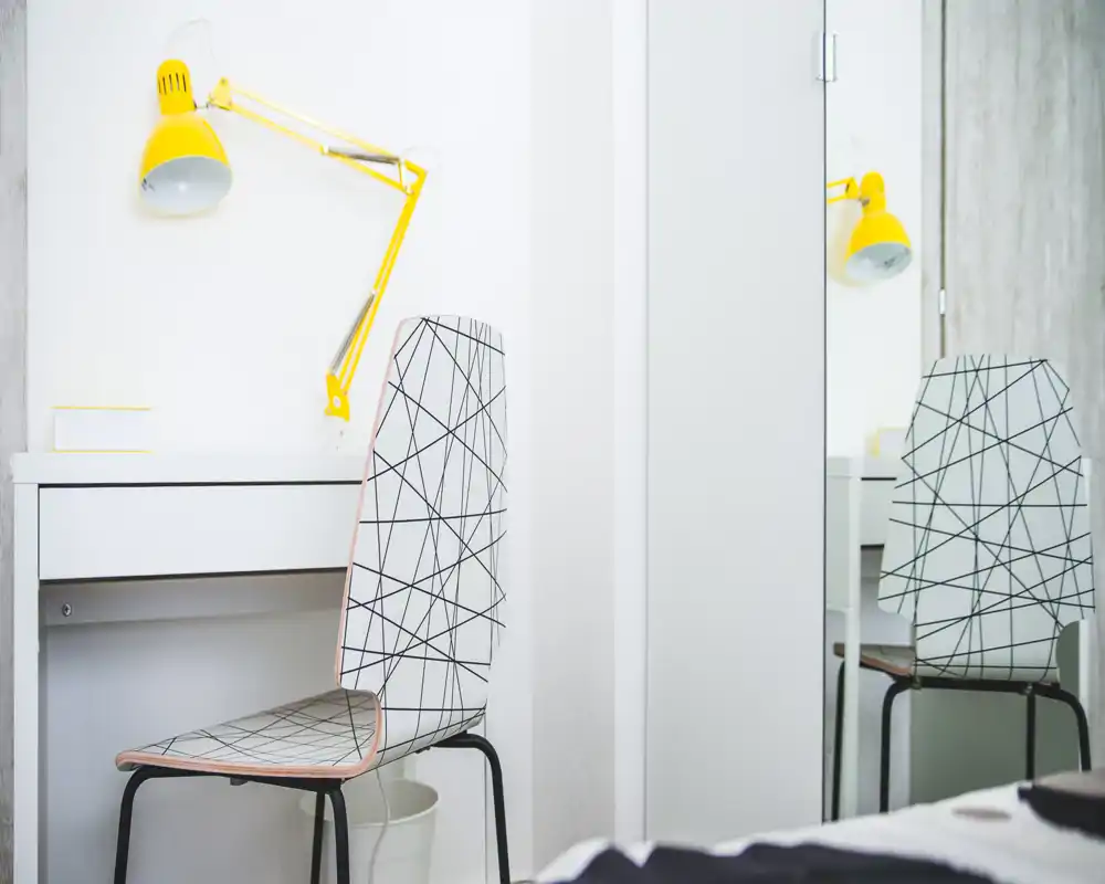 Minimalist bedroom corner with a patterned chair, small white desk and a bright yellow adjustable lamp; a small part of the bed is visible in the foreground. Clean, airy and well-lit space ideal for a study nook in the bedroom.
