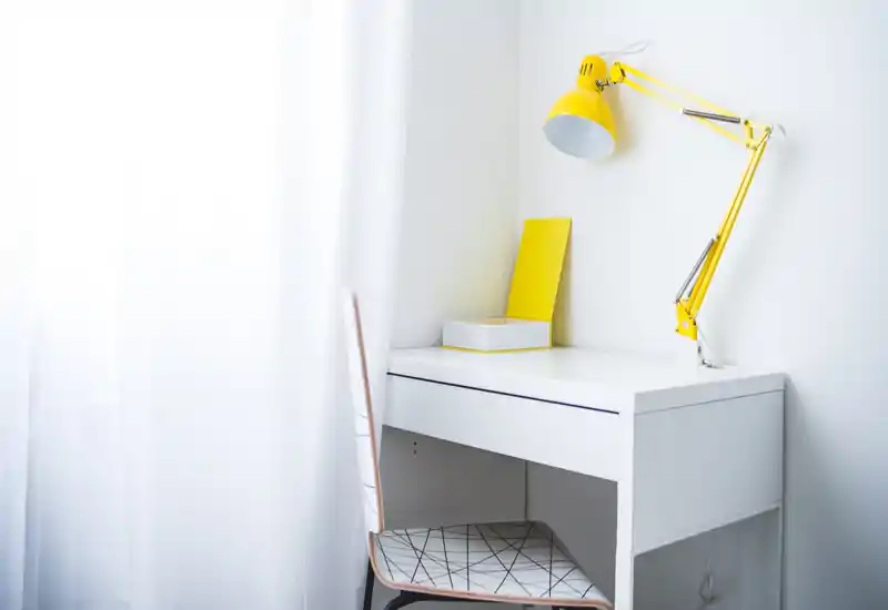 Bright, minimal corner of a bedroom featuring a white desk, patterned chair and a yellow wall lamp — simple, modern and airy.