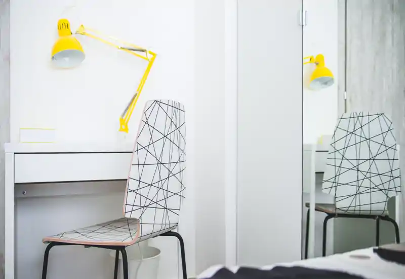 Minimalist bedroom corner with a patterned chair, small white desk and a bright yellow adjustable lamp; a small part of the bed is visible in the foreground. Clean, airy and well-lit space ideal for a study nook in the bedroom.
