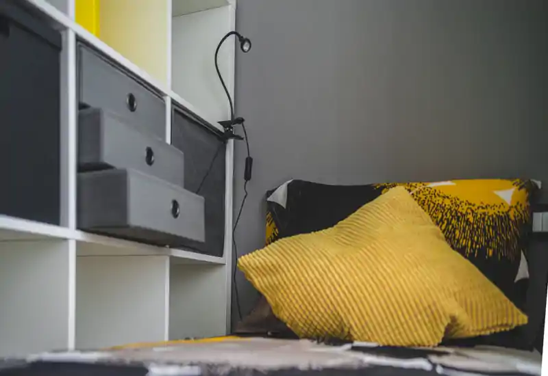 Cozy bedroom corner with textured yellow cushions and a neatly made bed beside open shelving; warm textiles and soft directional light create an inviting atmosphere.